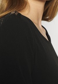 Czarny top z dekoltem w serek, zdobiony koronkowymi akcentami na ramionach, wykonany z miękkiego materiału o gładkiej fakturze.