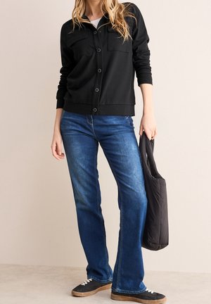 Frau, die eine schwarze, geknöpfte Jacke und blaue Jeans trägt, eine schwarze gesteppte Tragetasche hält und vor einem einfachen Hintergrund steht.