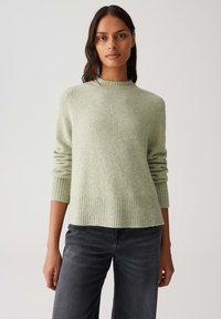 Woman with medium-dark skin and dark hair wears a light green knitted sweater and dark gray jeans, standing against a plain light background.