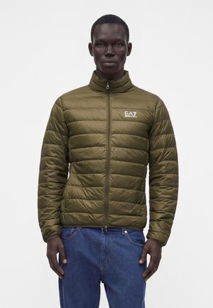 Man wearing olive green quilted jacket with EA7 logo on chest and blue jeans, standing against plain white background.