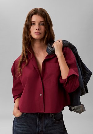 Jonge vrouw met lang bruin haar draagt een losse bordeauxrode blouse en donkere jeans, terwijl ze een donker spijkerjasje over haar schouder houdt tegen een grijze achtergrond.