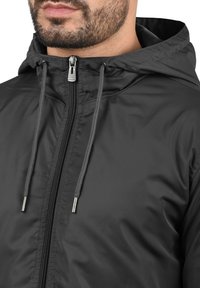 Svart huvad jacka med en slät, glänsande yta. Har en framdragkedja och dragsnören, med dragkedjor och detaljer i silverton.