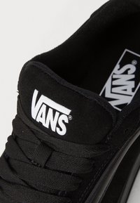 Gros plan sur la languette d'une chaussure Vans noire avec le logo Vans blanc et des lacets noirs sur fond blanc.