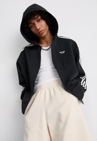 Sweatshirt preto com fecho éclair, com riscas brancas nas laterais e logótipo, combinado com calças bege de cintura elástica. Mistura de algodão, ajuste casual, tecido texturizado.