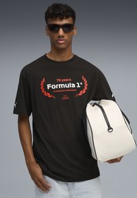 Sort t-shirt med røde og hvide "Formula 1" grafik, samt en hvid rygsæk med sorte detaljer og lynlås. Model bærer solbriller.