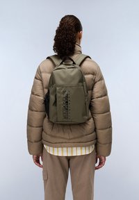 Olivgrüner Rucksack mit strukturiertem Finish, ausgestattet mit einer seitlichen Netzseitentasche, schwarzem Akzentlogo und verstellbaren Trägern. Trägt man über einer Daunenjacke.