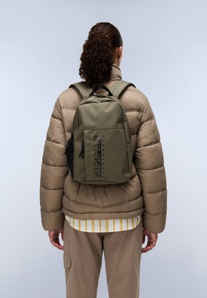 Mochila verde oliva con un acabado texturizado, que cuenta con un bolsillo de malla lateral, un logo de acento negro y correas ajustables. Llevada sobre una chaqueta acolchada.