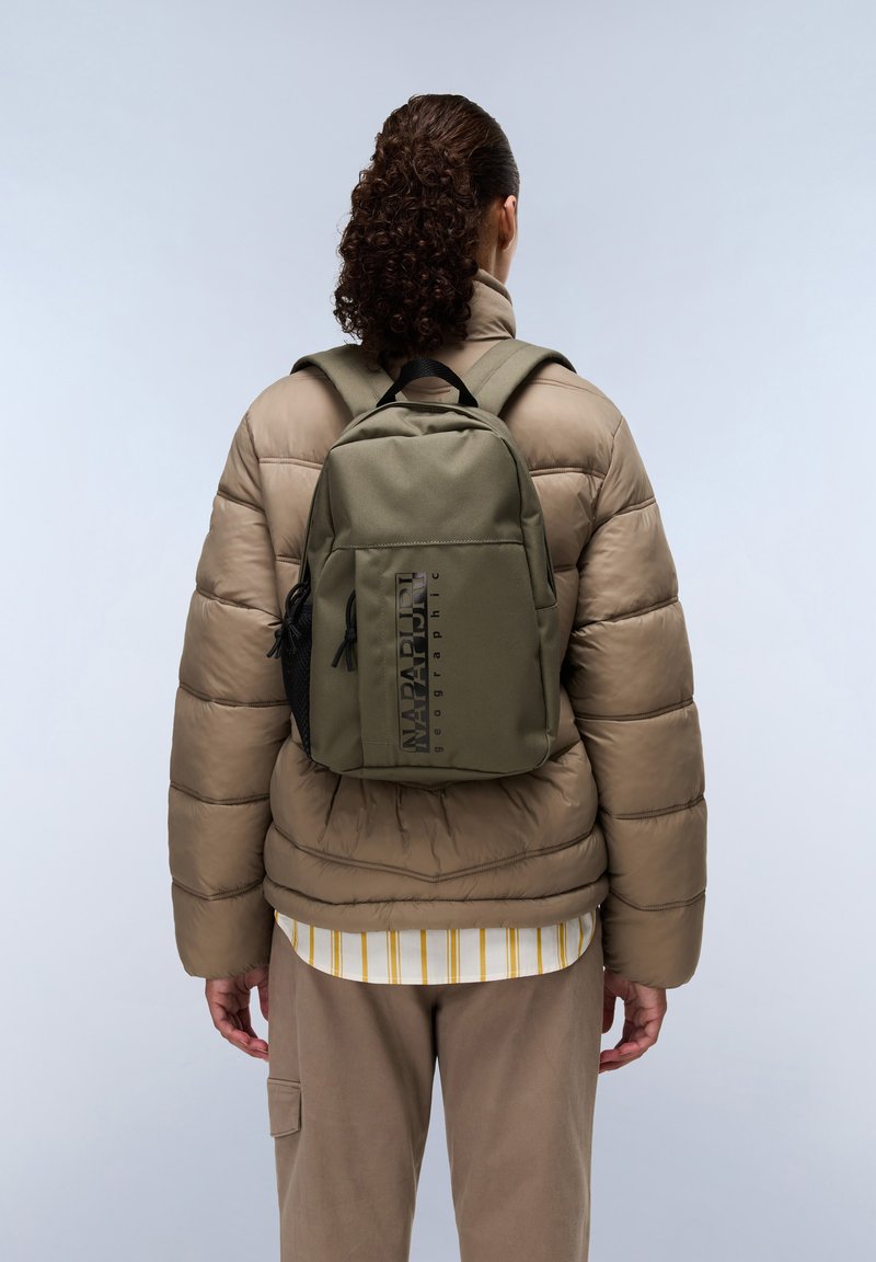 Olivgrüner Rucksack mit strukturiertem Finish, ausgestattet mit einer seitlichen Netzseitentasche, schwarzem Akzentlogo und verstellbaren Trägern. Trägt man über einer Daunenjacke.