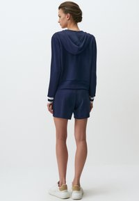 Set di felpa e pantaloni corti di colore navy, realizzato in tessuto morbido. Caratterizzato da un cappuccio, polsini a righe e abbinato a sneakers bianche.
