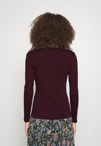 Kvinna med lockigt hår iklädd en långärmad mörklila tröja och en mörk blommig kjol, sedd bakifrån mot en enfärgad bakgrund.