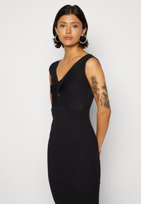 Vestido preto canelado com decote em V, design ajustado e detalhe torcido no busto. Sem mangas, com textura suave e silhueta justa.
