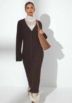Long, dark brown ribbed knitted cardigan with buttons, worn over a white high-neck top. Includes a tan shoulder bag and cream-colored pants.