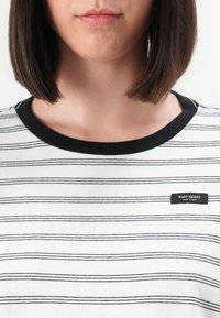 Žena s ravnom smešnom kosom nosi bijelu majicu s tankim crnim horizontalnim prugama i crnim izrezom s oznakom Kate Spade New York.