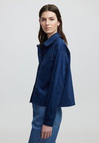Navy blauwe suède jas met een pointed collar, een enkele borstzak en drukknoopsluitingen, met een relaxte pasvorm en een textuurdetail op de zak.