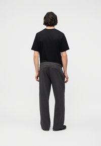 Άνδρας με σκούρα κυματιστά μαλλιά φοράει μαύρο πουκάμισο με κοντά μανίκια και άνετο σκουρόχρωμο γκρι παντελόνι, στέκεται κοιτώντας προς ένα απλό ανοιχτό γκρι φόντο.