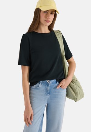 Kvinde iført gul kasket, sort t-shirt, lyseblå jeans, bærer en lysegrøn quiltet skuldertaske, står med den ene hånd i lommen.