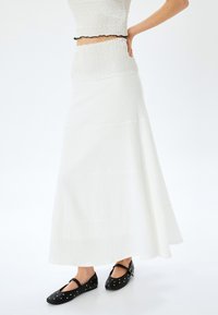 Jupe maxi blanche en étages avec une taille smockée. Le matériau semble léger avec une finition texturée. Associée à des chaussures plates noires ornées d'enclumes.