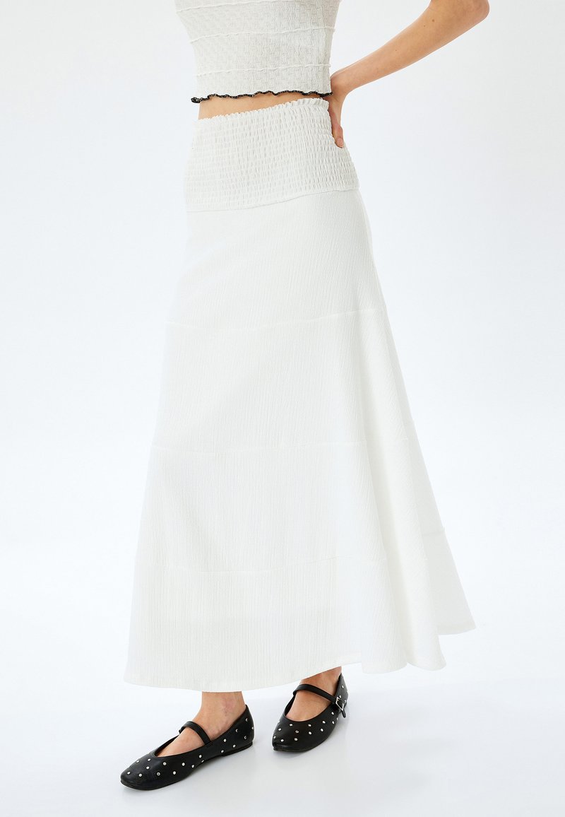 Jupe maxi blanche en étages avec une taille smockée. Le matériau semble léger avec une finition texturée. Associée à des chaussures plates noires ornées d'enclumes.