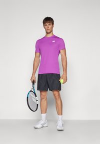 Camiseta de deporte corta de manga corta morada con logo blanco, combinada con pantalones cortos negros con estampado. Sostiene una raqueta de tenis y una pelota de tenis amarilla.