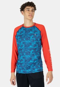 Langarmshirt mit blauem geometrischen Muster und roten Raglanärmeln. Hergestellt aus synthetischem Material, mit einem Logo auf der Brust.