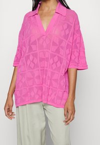 Rosa oversized polo shirt med ett texturerat geometriskt mönster, med korta ärmar och djup V-ringning. Lätt och andningsbart tyg.