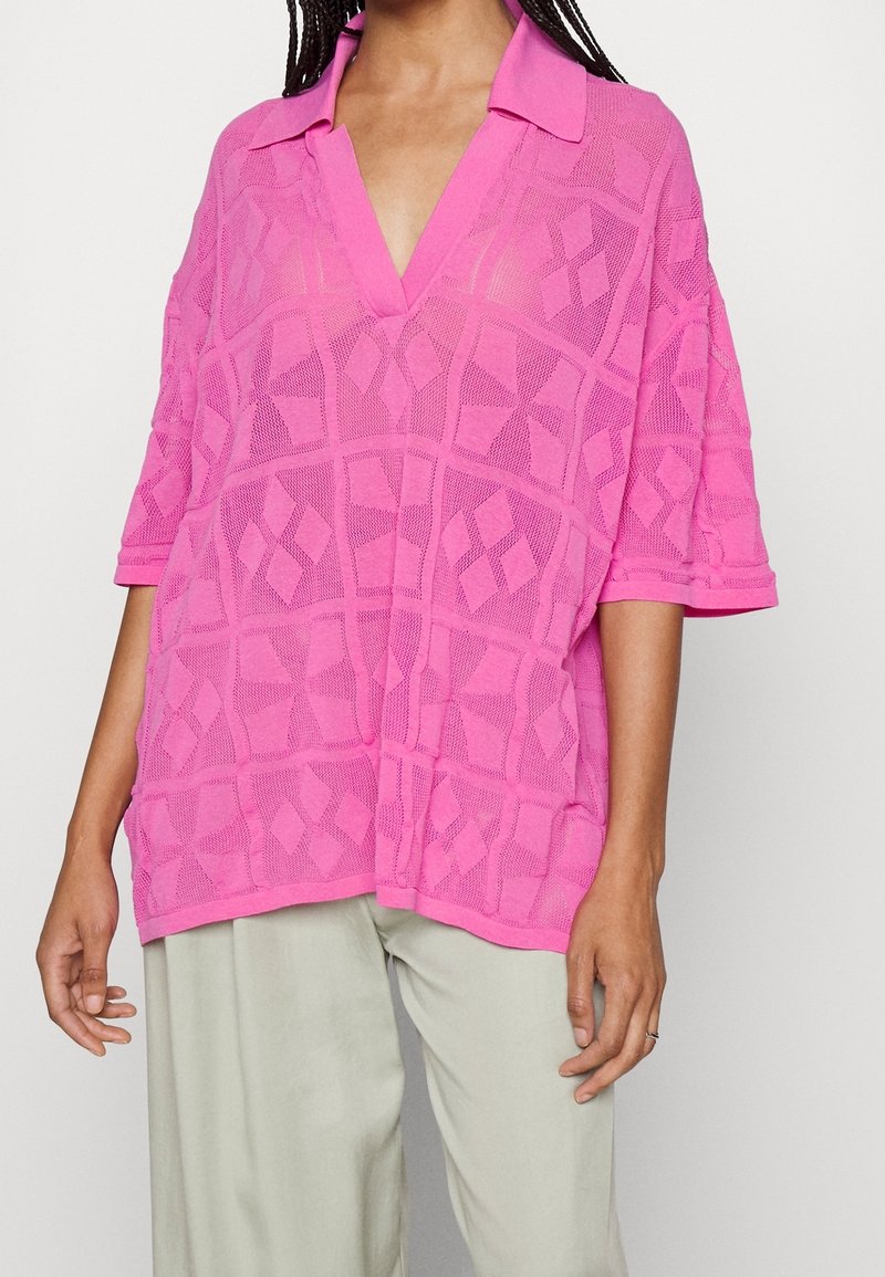 Rosa oversized polo shirt med ett texturerat geometriskt mönster, med korta ärmar och djup V-ringning. Lätt och andningsbart tyg.