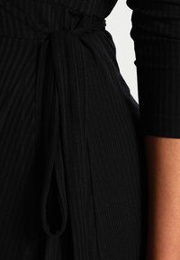 Svart ribbat tyg med midjeband. Designen har vertikala ränder och åtsittande långärmar som framhäver en mjuk textur.