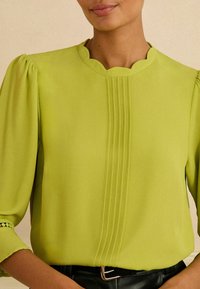 Limonkowa bluzka z falbaniastym dekoltem, plisami na środku i bufiastymi rękawami z dekoracyjnymi wycięciami na mankietach.