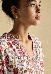 Hemdbluse aus Baumwolle mit floralem Muster in Weiß, versehen mit roten und grünen Blumen, einem V-Ausschnitt und kontrastierendem Stickerei am Kragen.