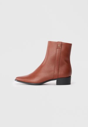 Brown leather ankle boot with low black heel, pointed toe, and small gold logo detail near the top on a white background.