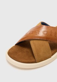 Sandale en daim marron et en cuir avec une large sangle embossée du mot "bugatti". Dispose d'une semelle en caoutchouc crème et d'une semelle intérieure rembourrée.