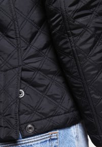 Sort quiltet jakke med diamantmønster, metallisk trykknaplukning og glat tekstur, parret med lyseblå denim.