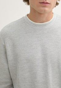 Grauer ribbeliger Pullover mit rundem Ausschnitt und subtiler Textur, über einem weißen Hemdkragen getragen. Einfaches, lässiges Design.