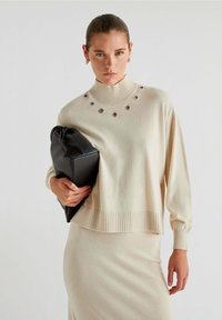 Beige striktrøje med ribbet krave og kant, med dekorative cirkulære udskæringer. Sættes sammen med en sort clutch.