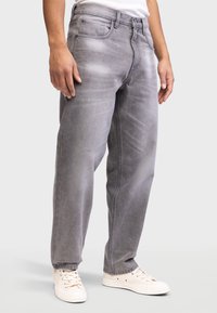 Svetlo sivi denim hlače s sproščenim krojem, ki imajo bledico, pet žepov in rahlo zožane hlačnice, v kombinaciji z belimi supergami.