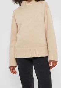 Pull à col roulé beige en matière tricotée, avec poignets et ourlet côtelés, au design simple et un petit détail de logo.