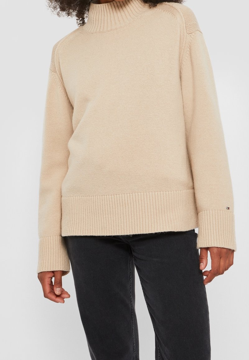 Pull à col roulé beige en matière tricotée, avec poignets et ourlet côtelés, au design simple et un petit détail de logo.