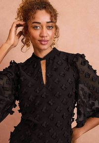 Rochie neagră cu modele florale texturate, mâneci din tul cu volane și decolteu cu tăietură. Purtați cu cercei aurii statement.