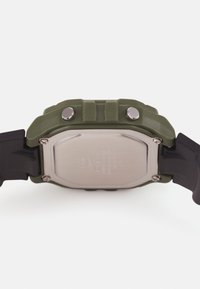 Montre en caoutchouc verte avec un dos en métal carré, dotée de deux boutons en métal argenté, d'un corps texturé et d'un bracelet noir, mettant en avant un design robuste.