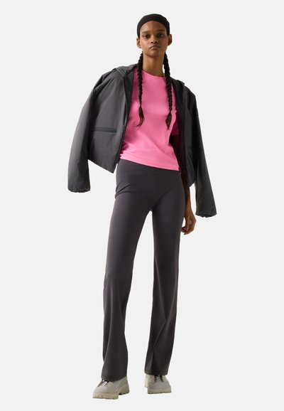 Junge Frau trägt eine schwarze Jacke über einem pinken Shirt, dunkle, eng anliegende Hose, weiße Turnschuhe und ein schwarzes Stirnband, steht vor einem einfarbigen Hintergrund.