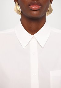 Lèvres brillantes, peau sombre, portant de grandes boucles d'oreilles abstraites en or et une chemise blanche à col boutonnée avec une poche.