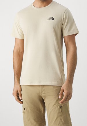 Hombre vistiendo una camiseta de manga corta beige con el logo de The North Face y pantalones cortos cargo caqui con bolsillos con cremallera, de pie contra un fondo blanco.
