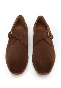 Zapatos de ante marrón con textura suave, que presentan una correa cruzada y detalle de hebilla. Punta redondeada y tacón bajo. Diseño clásico.