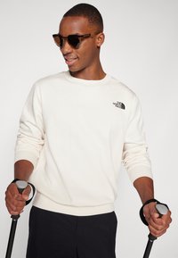 Creme sweatshirt med ribbede manchetter og kant, prydet med et sort logo på brystet. Sorte bukser og solbriller fuldender outfittet.