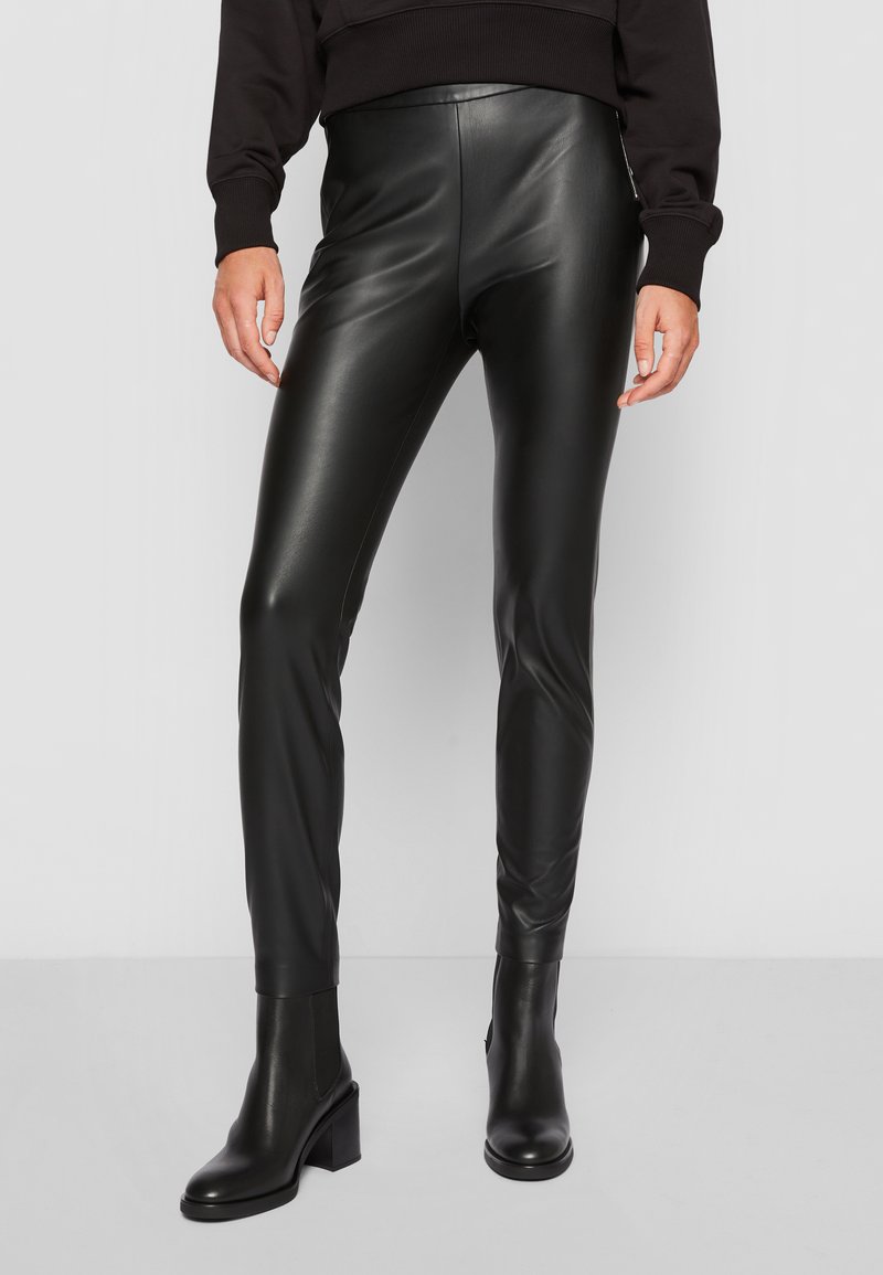 Hoge taille zwarte faux leren leggings met een gladde textuur, een slanke pasvorm en minimale stikseldetails, gecombineerd met enkellaarsjes met een gestapelde hak.