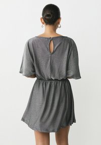 Rochie metalizată gri cu mâneci scurte, talie elastică și detaliu de cheiță la spate. Textură netedă cu un finisaj strălucitor.