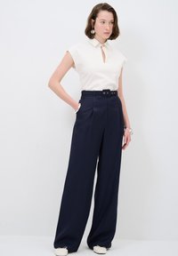 Un haut crème avec un col et un détail en trou de serrure, associé à un pantalon large taille haute bleu marine et des chaussures de couleur claire.