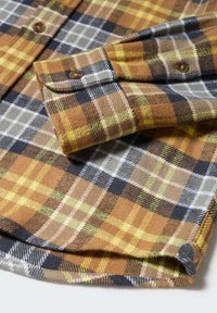 Manche de chemise en flanelle à carreaux marron, jaune, gris et noir avec poignet boutonné sur une surface blanche.