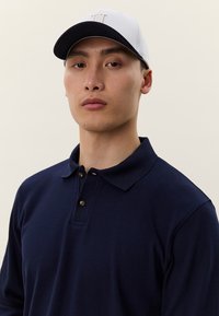 Ung mann med hvit og svart baseballcap og marineblå langermet polo, ser rett frem mot en ensfarget bakgrunn.