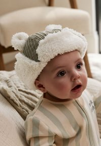 Chlpatá biela a olivovo zelená čiapka s brmbolcami, doplnená pruhovaným béžovým a svetlozeleným overalom. Textúrované tkaniny vytvárajú útulný vzhľad.